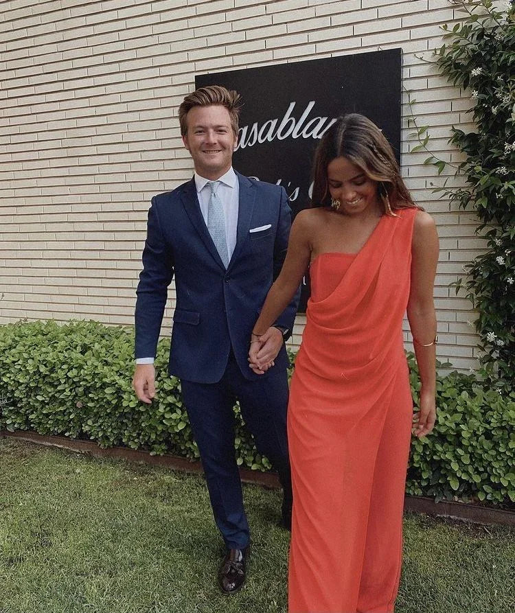 Man in navy suit and woman in red one-shoulder dress holding hands outdoors near ZAPDRESS sign.