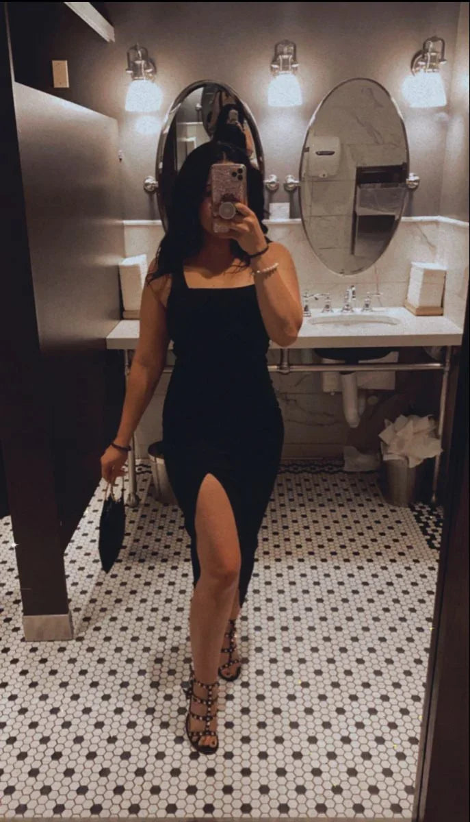 Woman in black sleeveless dress with slit taking mirror selfie in modern restroom