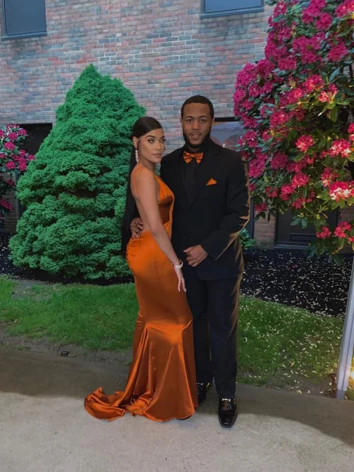 Couple posing outdoors, woman in elegant orange satin gown, man in black suit with orange accents, lush greenery and blooming flowers, ZAPDRESS style