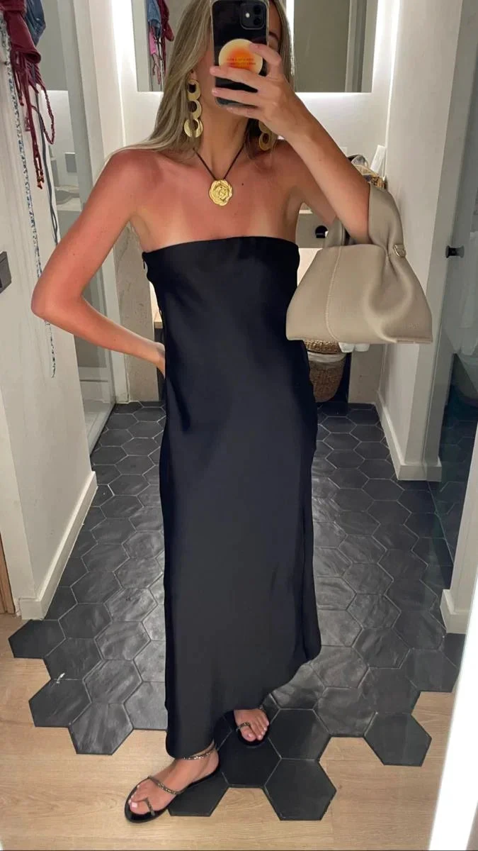 Woman wearing a black strapless dress accessorized with gold jewelry and a beige handbag, standing in a modern room, ZAPDRESS fashion.