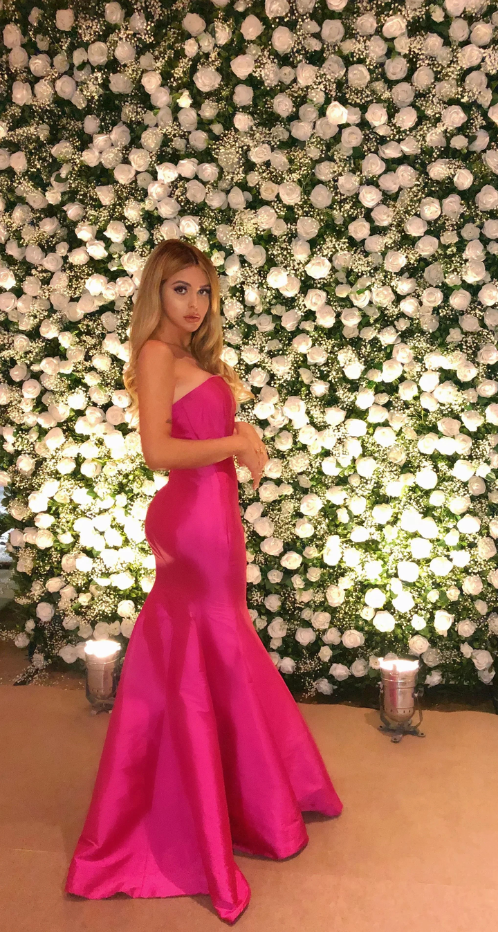 Woman in bright pink evening gown posing by white rose flower wall at ZAPDRESS event