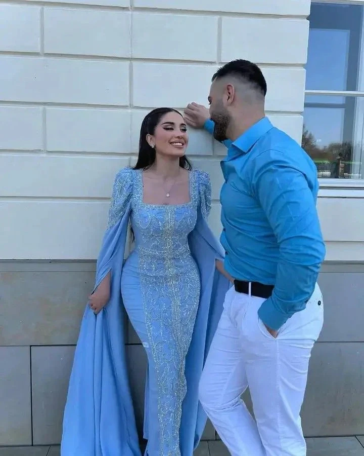 Woman in shimmering blue formal dress and man in blue shirt posing by white wall, ZAPDRESS style