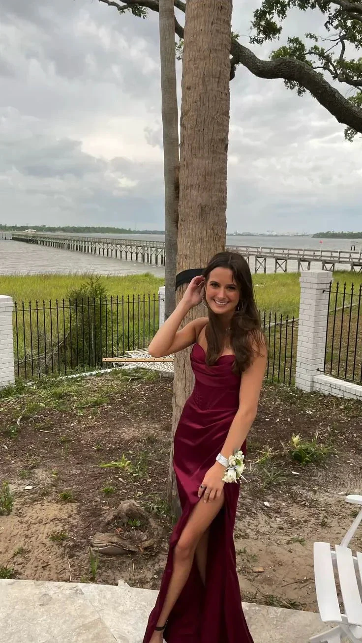 Smiling woman in elegant burgundy evening dress standing outdoors near waterfront and pier