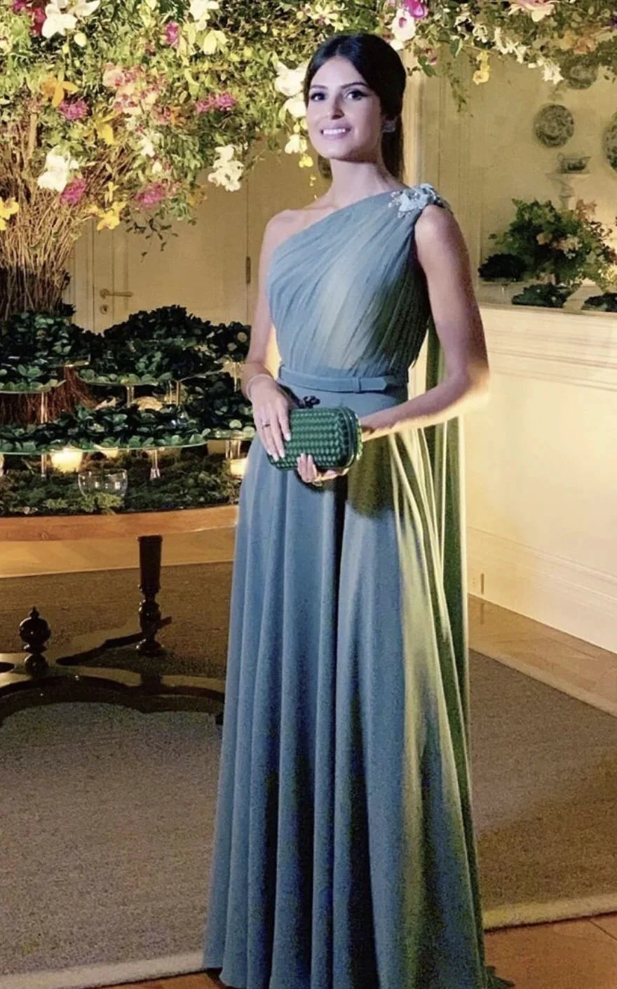 Woman in elegant one-shoulder blue evening gown holding a green clutch at a floral event